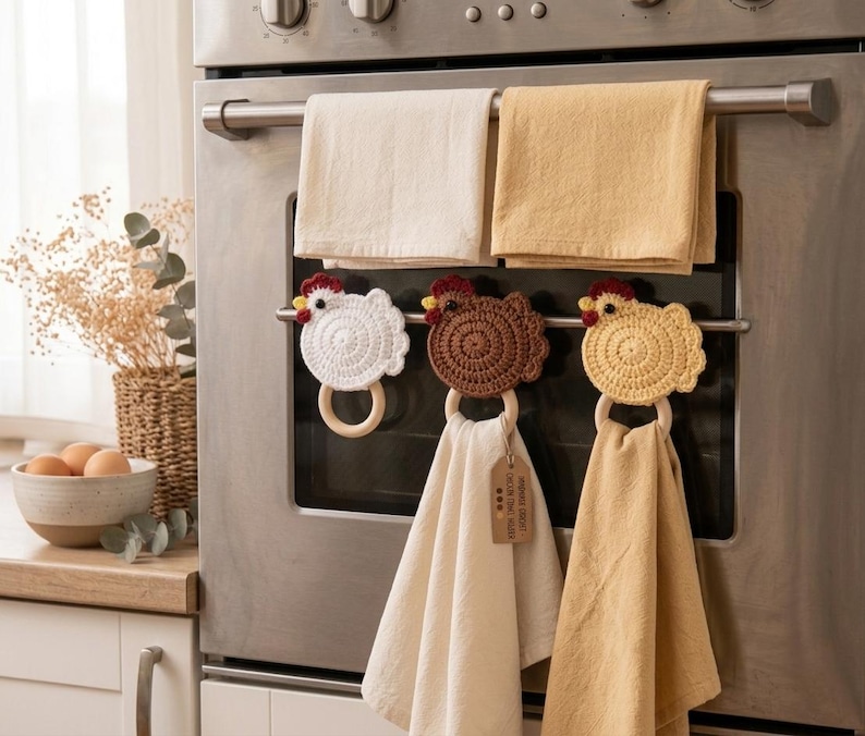 May include: Three crochet chicken towel holders in white, brown, and yellow, each featuring a wooden ring. Two plain rectangular kitchen towels hang from the oven handle, while two additional towels are suspended from the chicken-shaped holders.