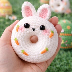 May include: A handmade white crochet donut with bunny ears and a face, held in a hand. The donut has black eyes, pink cheeks, and carrot decorations. Other Easter-themed crochet items are in the background.