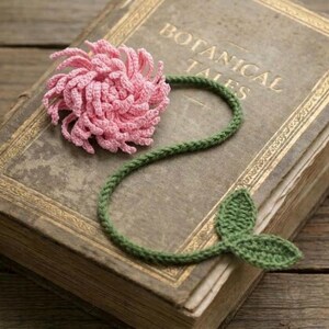 Può includere: Segnalibro all'uncinetto fatto a mano con un fiore rosa e uno stelo verde con foglie, appoggiato su un libro antico intitolato "Botanical Tales". Il libro ha bordi dorati ed è posto su una superficie di legno.