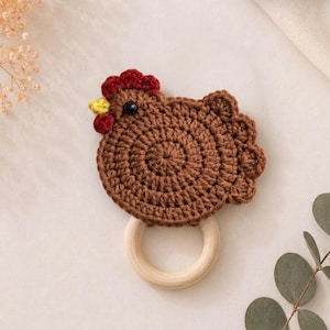 May include: A brown crocheted chicken toy attached to a smooth, round, light-colored wooden ring. The chicken features a dark red comb and wattle, a small yellow beak, and a single black bead eye, with a textured, circular yarn body design.
