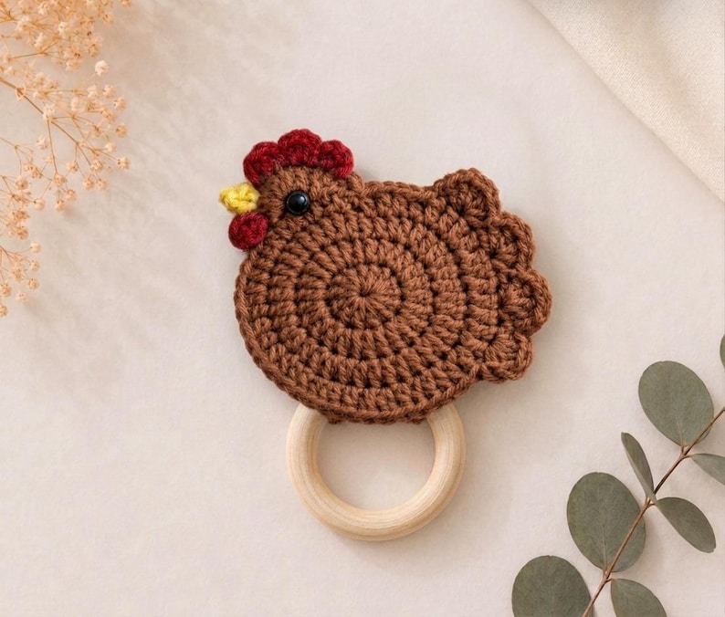 May include: A brown crocheted chicken toy attached to a smooth, round, light-colored wooden ring. The chicken features a dark red comb and wattle, a small yellow beak, and a single black bead eye, with a textured, circular yarn body design.