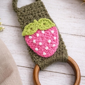 Op de afbeelding: Een handgemaakte gehaakte handdoekhouder met een roze aardbei met witte stippen en groene bladeren. De houder is olijfgroen met een houten ring en lus om op te hangen. De aardbei is in het midden geplaatst.