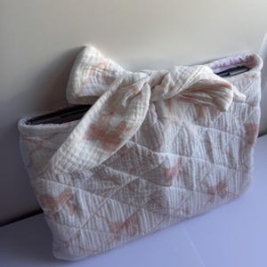May include: A quilted, off-white clutch with a bow closure. The clutch features a diamond pattern and a delicate pink floral print. The bow is made of the same fabric. The interior is visible.