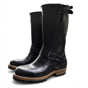 May include: A pair of black leather mid-calf boots featuring a rounded toe, a thick lugged sole with a visible light brown wooden midsole, and two adjustable straps with metal buckles located at the ankle and the top outer shaft of the boots.