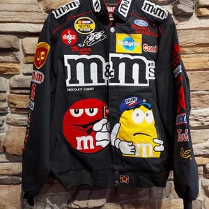Peut inclure: Veste de course noire M&M's avec de nombreux écussons et logos, dont M&M's et NASCAR. La veste a une fermeture boutonnée et un col. Elle présente des accents rouges, blancs, jaunes et bleus.