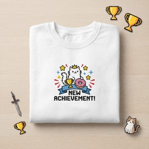 May include: A white t-shirt featuring a graphic of a white cat wearing a crown, holding a gold trophy and a pink doughnut. Above the cat are yellow stars and blue sparkles. Below the cat is a blue banner with the text NEW ACHIEVEMENT! in black.