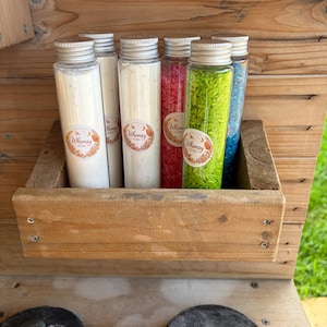 May include: Six clear plastic tubes filled with colourful grains, including white, pink, green, and blue, are arranged in a wooden crate. Each tube features a round label with the text Whimsy Kids and a silver screw-top lid.
