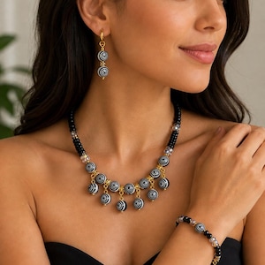 May include: A black and white beaded jewellery set, including a necklace, bracelet, and earrings. The necklace and earrings feature dangling spiral-patterned beads. The bracelet has a mix of black and clear beads. Gold-coloured accents are present throughout the set.