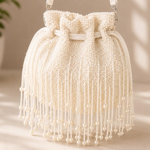 Può includere: Una borsa a secchiello color crema con coulisse, ricoperta di piccole perline. La borsa presenta lunghe frange di perline pendenti che terminano con perline rotonde più grandi. Include una spessa corda intrecciata e una tracolla abbinata.