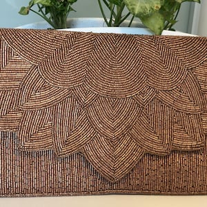 Puede incluir: Bolso de mano rectangular con cuentas de color bronce con cierre de solapa. La parte delantera presenta un patrón detallado en forma de abanico en un tono bronce más claro, creando un diseño decorativo texturizado. Un accesorio elegante.