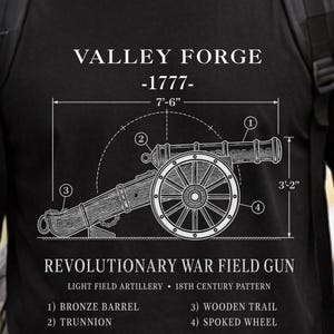 May include: A black t-shirt featuring a white technical diagram of a Valley Forge 1777 Revolutionary War field gun. The print includes labels for the bronze barrel, trunnion, wooden trail, and spoked wheel, with dimensions of 7'-6" and 3'-2".