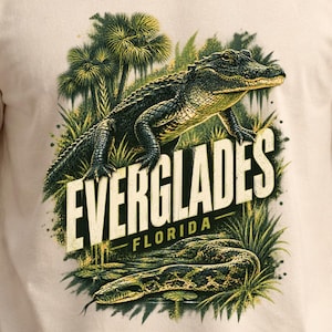 May include: A beige short-sleeved t-shirt featuring a graphic print of an alligator in a swampy landscape with palm trees and tall grass. The text "EVERGLADES" is printed in large cream letters above the word "FLORIDA" in smaller text.