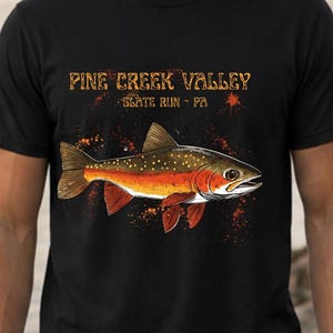 May include: Black t-shirt featuring a detailed illustration of a fish with orange, yellow, and red accents. The shirt has the text "PINE CREEK VALLEY - SLATE RUN - PA" in a distressed font, with orange paint splatter details.