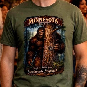 May include: Olive green t-shirt featuring a graphic of a Bigfoot figure peeking from behind a tree. The graphic includes the text "MINNESOTA Est. 1858" and "Northwoods Sasquatch". The shirt is made of a soft material.