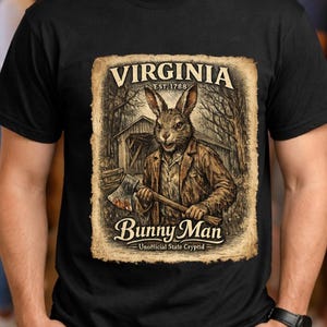 May include: Black t-shirt featuring a vintage-style graphic. The graphic depicts a rabbit-like figure in a brown coat holding an axe, with the text "VIRGINIA EST. 1788" and "Bunny Man - Unofficial State Cryptid".