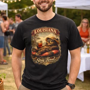May include: Black t-shirt with a graphic of a bear in a straw hat, sunglasses, and an inner tube, holding a drink. The shirt reads "LOUISIANA Est. 1812" above the bear and "Stay Feral" below. The person is wearing khaki pants.