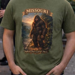 May include: Olive green t-shirt featuring a graphic of a Bigfoot-like creature in a forest setting, with the text "MISSOURI" and "Unofficial State Cryptid: Momo". The shirt has a crew neck and short sleeves.