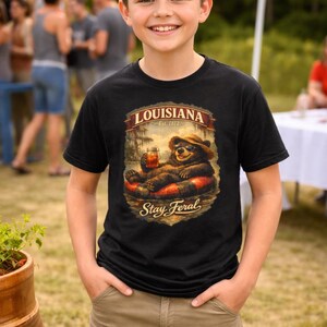May include: Black t-shirt with a graphic featuring a bear in a hat and sunglasses, relaxing on an inner tube with a drink. The shirt has the text "LOUISIANA Est. 1812" and "Stay Feral". The boy is wearing khaki shorts.
