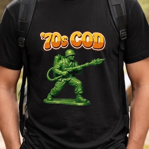 May include: Black t-shirt featuring a green toy soldier with a flamethrower. The shirt has the text "70s COD" in orange and yellow. The toy soldier is a classic design, reminiscent of vintage toys.