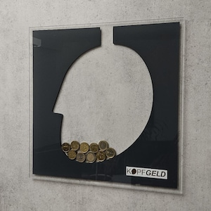 Op de afbeelding: Een ingelijst kunstwerk met een grijze, betonachtige rand. Binnenin is een zwart silhouet van een hoofdvorm te zien, met een verzameling munten aan de onderkant. Het woord "KOPFGELD" is gedrukt op een witte sticker.
