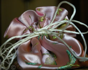 May include: A pink satin drawstring bag with a white cord. The bag has a floral pattern with green and gold accents.