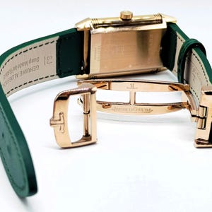 Puede incluir: Un reloj rectangular dorado con una correa de cuero verde. El reloj tiene un cierre dorado y las palabras "JAEGER-LECOULTRE" son visibles en el cierre. La correa est&aacute; abierta, revelando la parte inferior del reloj.
