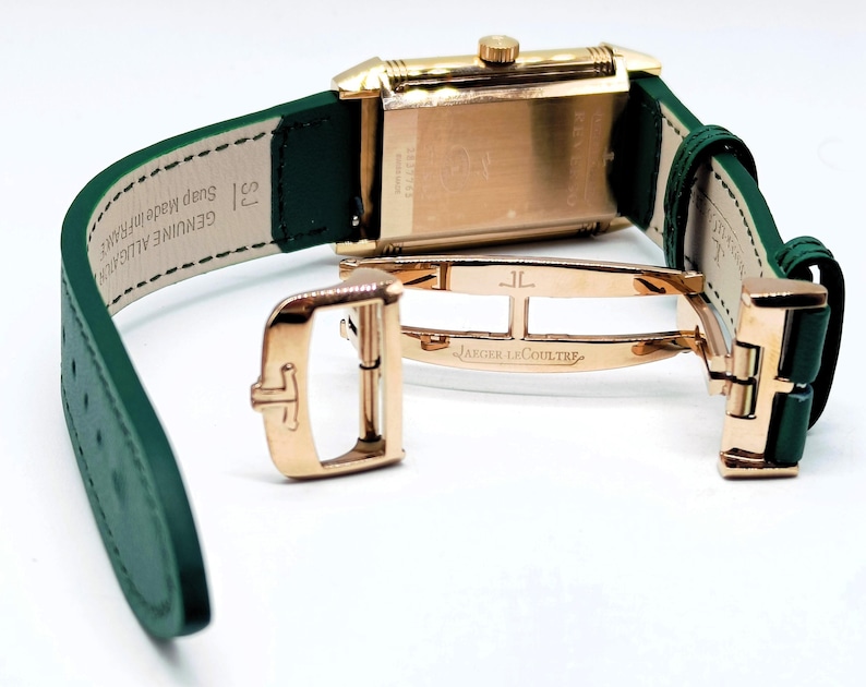 Puede incluir: Un reloj rectangular dorado con una correa de cuero verde. El reloj tiene un cierre dorado y las palabras "JAEGER-LECOULTRE" son visibles en el cierre. La correa est&aacute; abierta, revelando la parte inferior del reloj.