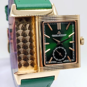 Puede incluir: Un reloj de pulsera rectangular dorado con esfera verde y un peque&ntilde;o dial de segundos. El reloj tiene una correa de cuero verde. La marca "JAEGER-LECOULTRE" es visible en la esfera. El reloj est&aacute; marcado "SWISS MADE".