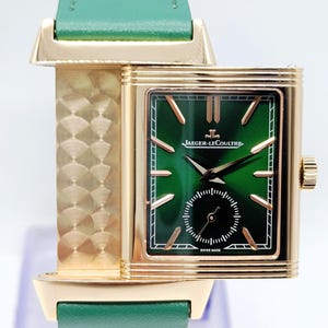 Puede incluir: Un reloj Jaeger-LeCoultre dorado con esfera verde y correa de cuero verde. El reloj tiene una esfera rectangular con agujas e indicadores dorados. La correa mide aproximadamente 2 cm de ancho.