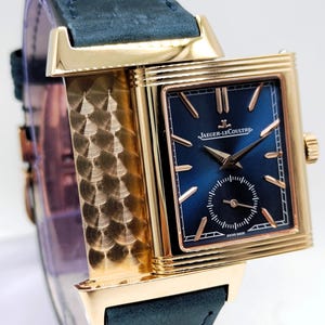 K&ouml;nnte beinhalten: Eine goldfarbene Jaeger-LeCoultre-Uhr mit dunkelblauem Zifferblatt und kleiner Sekunde. Die Uhr hat goldene Zeiger und Markierungen, ein rechteckiges Geh&auml;use und ein dunkelblaues Lederarmband. Auf dem Zifferblatt steht "JAEGER-LECOULTRE" und "SWISS MADE".