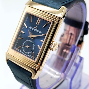 K&ouml;nnte beinhalten: Eine rechteckige Armbanduhr in Goldoptik mit einem marineblauen Zifferblatt und einem dunkelblauen Lederarmband. Das Zifferblatt der Uhr hat goldfarbene Zeiger und Markierungen, mit dem Markennamen "Jaeger-LeCoultre". Die Uhr wird auf einem transparenten St&auml;nder pr&auml;sentiert.