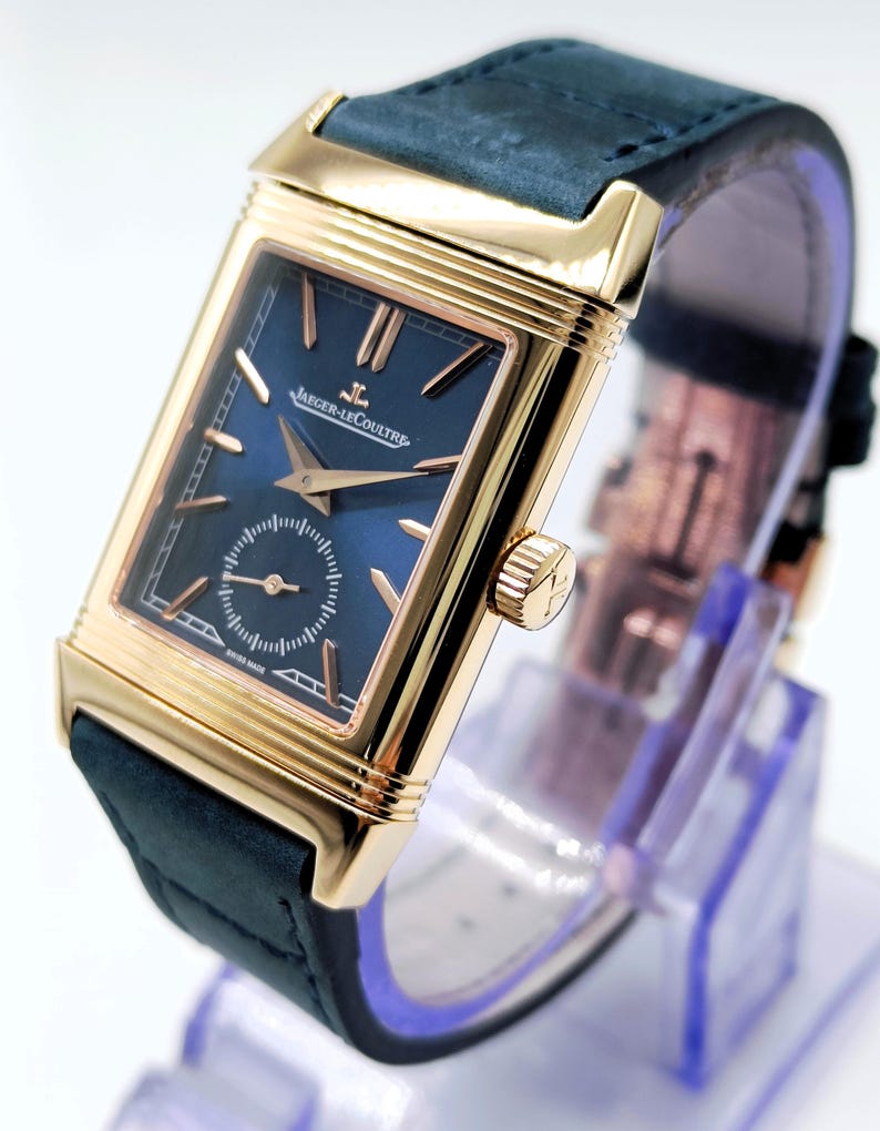 K&ouml;nnte beinhalten: Eine rechteckige Armbanduhr in Goldoptik mit einem marineblauen Zifferblatt und einem dunkelblauen Lederarmband. Das Zifferblatt der Uhr hat goldfarbene Zeiger und Markierungen, mit dem Markennamen "Jaeger-LeCoultre". Die Uhr wird auf einem transparenten St&auml;nder pr&auml;sentiert.