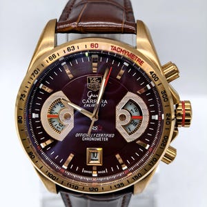Op de afbeelding: Een TAG Heuer Grand Carrera Calibre 17 RS2 polshorloge met een goudkleurige kast, een diepbordeauxrode wijzerplaat en een bruine lederen band. De wijzerplaat is voorzien van een tachymeterschaal, twee sub-wijzerplaten, een datumvenster en tekst.