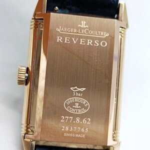 K&ouml;nnte beinhalten: Eine goldfarbene Jaeger-LeCoultre Reverso Uhr mit rechteckigem Zifferblatt und dunkelblauem Lederarmband. Das Zifferblatt zeigt den Markennamen und das Modell sowie den Text "3 bar", "1000 Hours Control" und die Seriennummer 277.8.62.