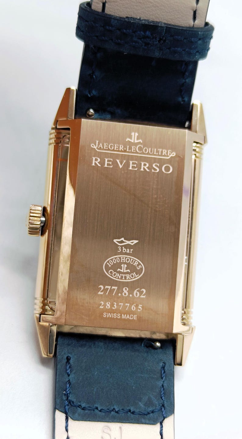 K&ouml;nnte beinhalten: Eine goldfarbene Jaeger-LeCoultre Reverso Uhr mit rechteckigem Zifferblatt und dunkelblauem Lederarmband. Das Zifferblatt zeigt den Markennamen und das Modell sowie den Text "3 bar", "1000 Hours Control" und die Seriennummer 277.8.62.