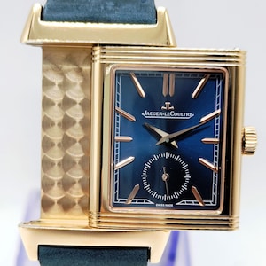 K&ouml;nnte beinhalten: Eine rechteckige Armbanduhr in Gold mit einem marineblauen Zifferblatt und einer kleinen Sekundenscheibe. Die Uhr hat goldene Zeiger und Markierungen, ein marineblaues Lederarmband und das Jaeger-LeCoultre-Logo. Die Uhr ist mit "Swiss Made" gekennzeichnet.