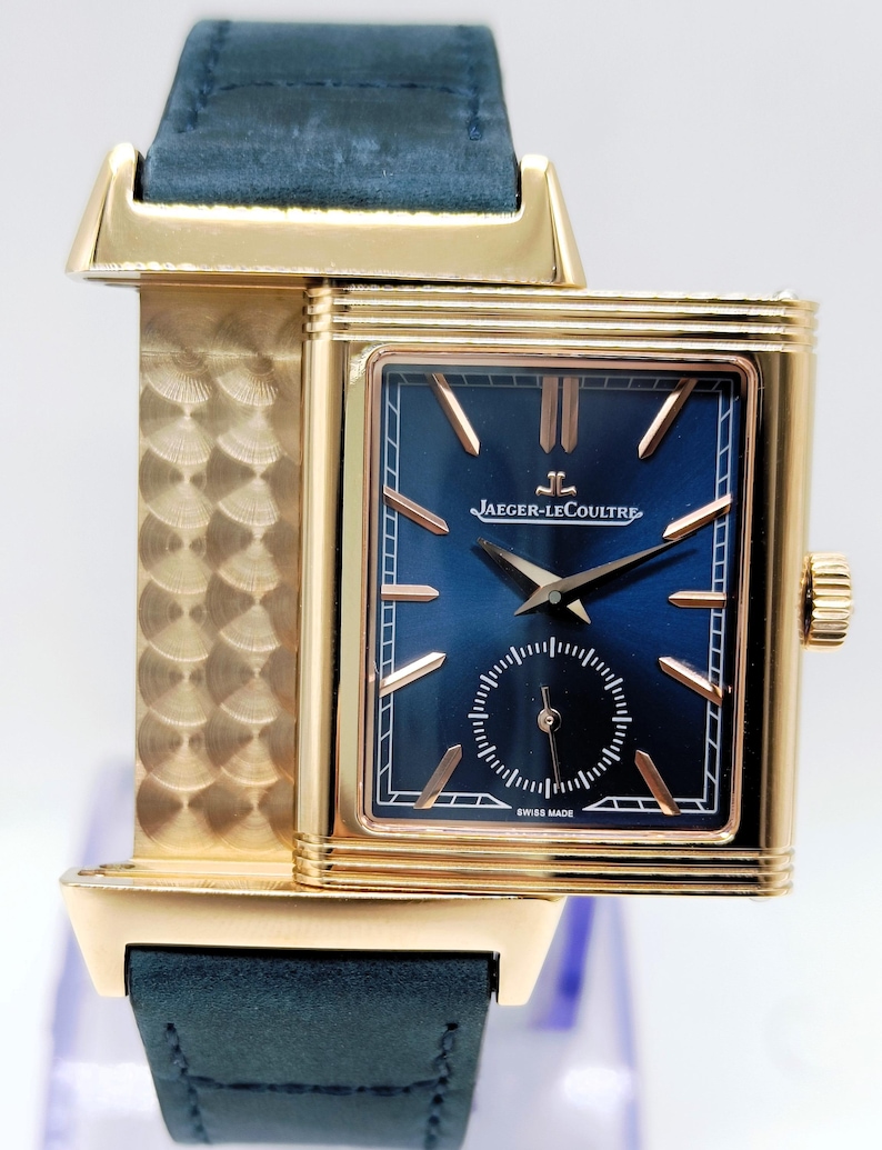 K&ouml;nnte beinhalten: Eine rechteckige Armbanduhr in Gold mit einem marineblauen Zifferblatt und einer kleinen Sekundenscheibe. Die Uhr hat goldene Zeiger und Markierungen, ein marineblaues Lederarmband und das Jaeger-LeCoultre-Logo. Die Uhr ist mit "Swiss Made" gekennzeichnet.