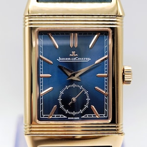 K&ouml;nnte beinhalten: Eine rechteckige Armbanduhr in Goldoptik mit blauem Zifferblatt und einem marineblauen Lederarmband. Das Zifferblatt der Uhr hat goldfarbene Zeiger und Markierungen mit dem Markennamen "JAEGER-LECOULTRE". Die Uhr ist mit "SWISS MADE" gekennzeichnet.