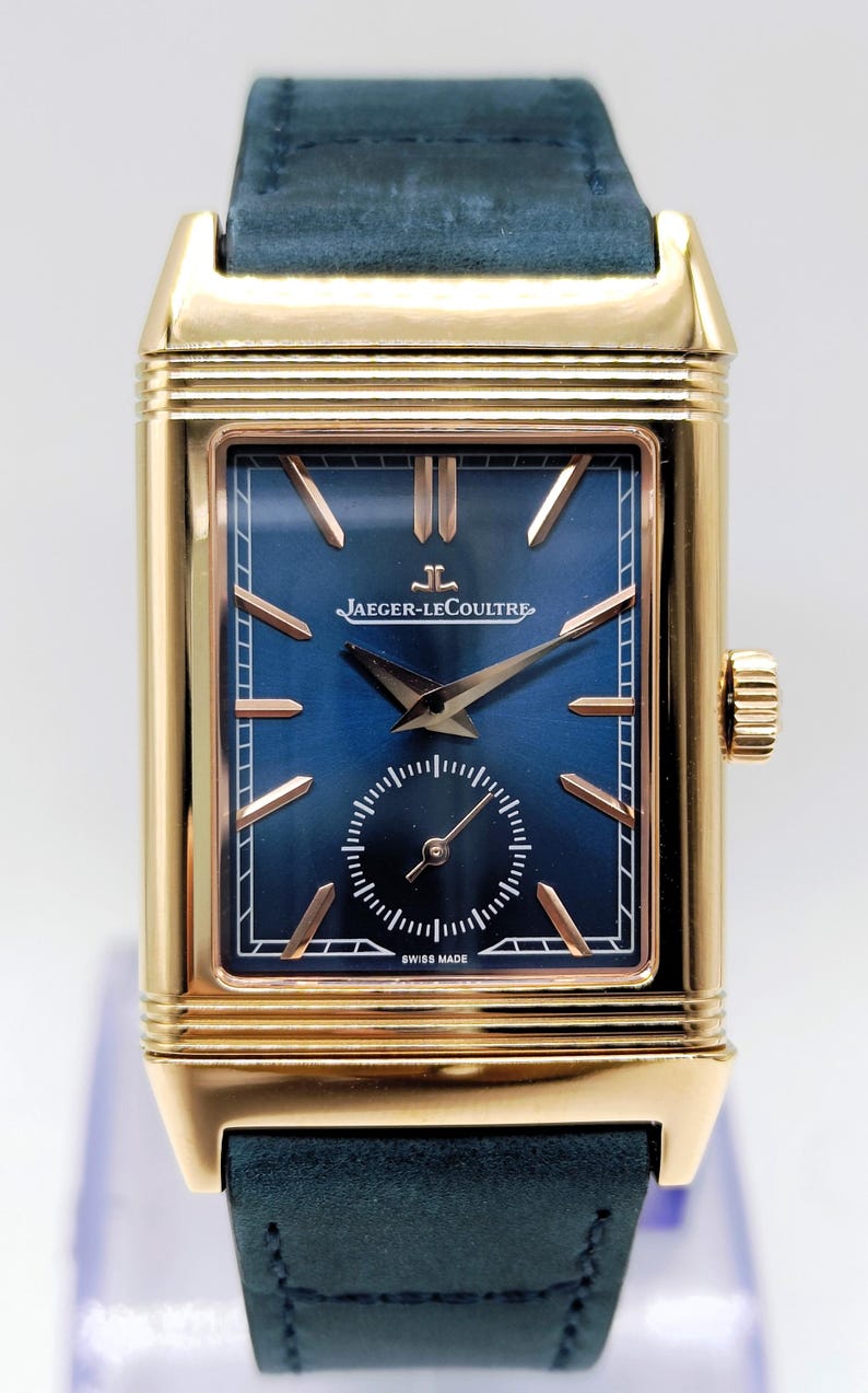 K&ouml;nnte beinhalten: Eine rechteckige Armbanduhr in Goldoptik mit blauem Zifferblatt und einem marineblauen Lederarmband. Das Zifferblatt der Uhr hat goldfarbene Zeiger und Markierungen mit dem Markennamen "JAEGER-LECOULTRE". Die Uhr ist mit "SWISS MADE" gekennzeichnet.