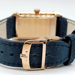 K&ouml;nnte beinhalten: Nahaufnahme einer Luxus-Armbanduhr. Die Uhr hat ein goldfarbenes rechteckiges Geh&auml;use und ein marineblaues Lederarmband. Die Schnalle und der Verschluss sind ebenfalls goldfarben. Die Uhr ist mit einer klassischen und eleganten &Auml;sthetik gestaltet.