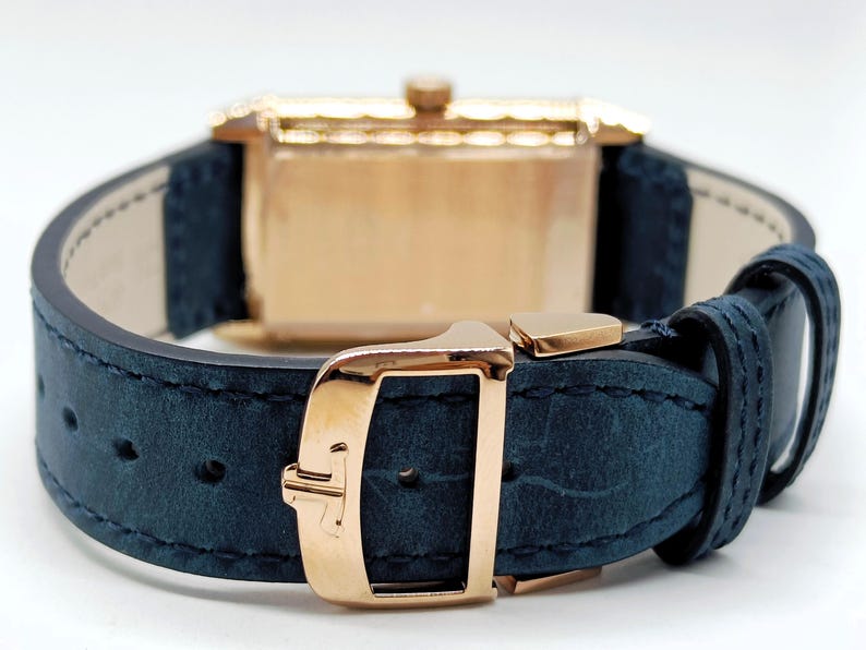 K&ouml;nnte beinhalten: Nahaufnahme einer Luxus-Armbanduhr. Die Uhr hat ein goldfarbenes rechteckiges Geh&auml;use und ein marineblaues Lederarmband. Die Schnalle und der Verschluss sind ebenfalls goldfarben. Die Uhr ist mit einer klassischen und eleganten &Auml;sthetik gestaltet.