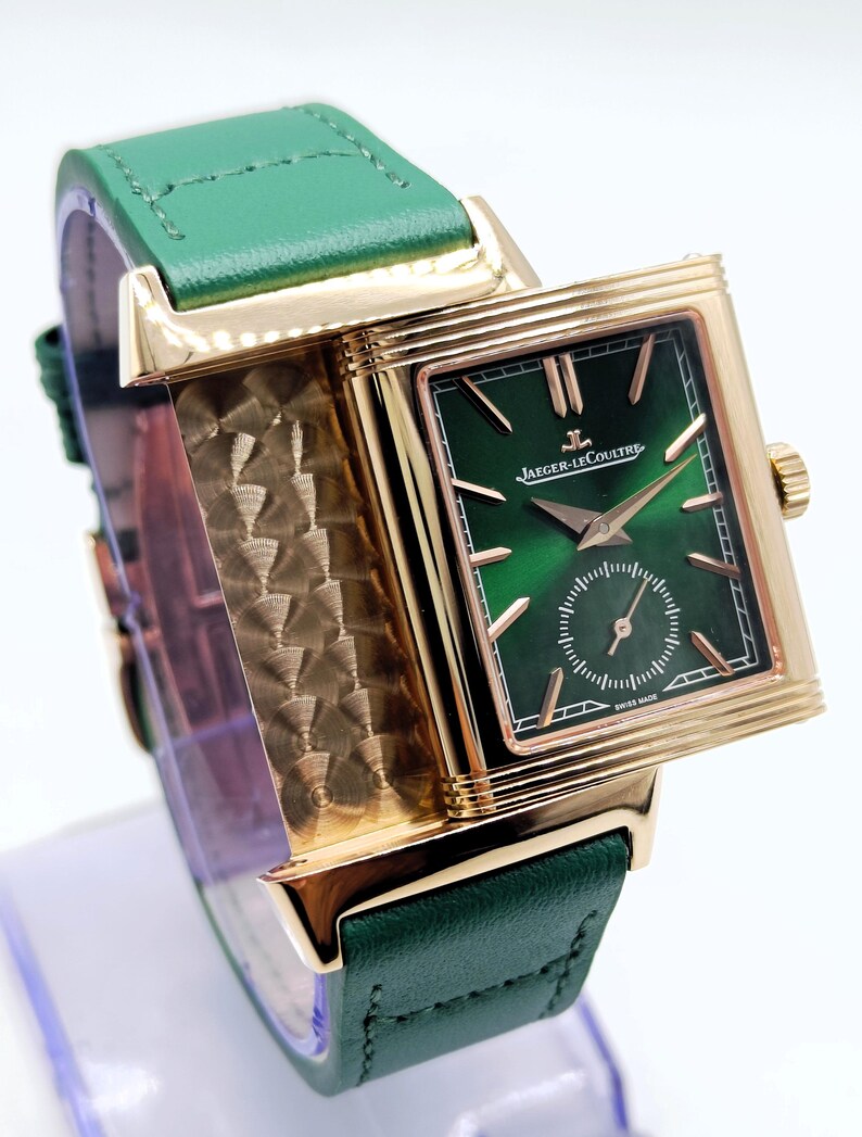 Puede incluir: Un reloj rectangular dorado con esfera verde y correa de cuero verde. La esfera del reloj muestra el texto "Jaeger-LeCoultre" y "Swiss Made". El reloj tiene un peque&ntilde;o dial de segundos y agujas doradas.