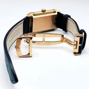 K&ouml;nnte beinhalten: Eine rechteckige Armbanduhr in Gold mit einem schwarzen Lederarmband. Die Uhr hat einen goldenen Verschluss mit der Aufschrift "JAEGER-LECOULTRE". Die Uhr wird vor einem wei&szlig;en Hintergrund pr&auml;sentiert, um ihr Design und ihre Handwerkskunst hervorzuheben.