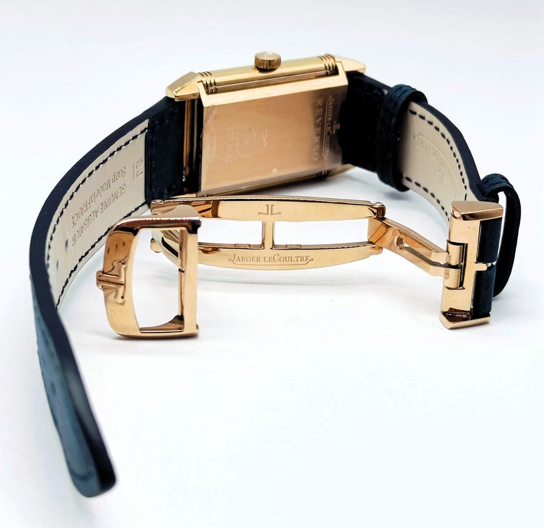 K&ouml;nnte beinhalten: Eine rechteckige Armbanduhr in Gold mit einem schwarzen Lederarmband. Die Uhr hat einen goldenen Verschluss mit der Aufschrift "JAEGER-LECOULTRE". Die Uhr wird vor einem wei&szlig;en Hintergrund pr&auml;sentiert, um ihr Design und ihre Handwerkskunst hervorzuheben.