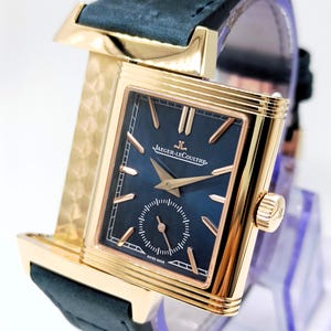 K&ouml;nnte beinhalten: Eine goldfarbene rechteckige Armbanduhr mit dunkelblauem Zifferblatt und kleiner Sekunde. Die Uhr hat goldene Zeiger und Markierungen, ein schwarzes Lederarmband und den Text "JAEGER-LECOULTRE SWISS MADE".
