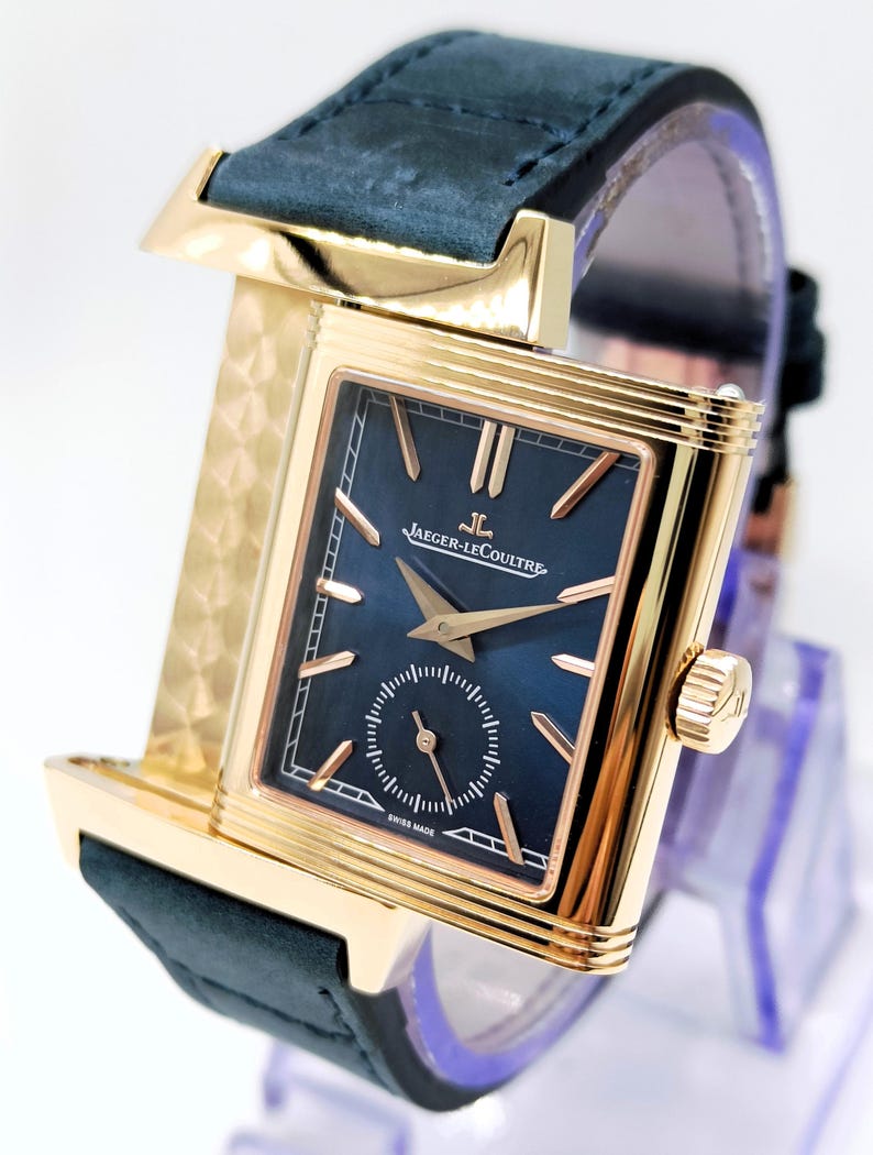 K&ouml;nnte beinhalten: Eine goldfarbene rechteckige Armbanduhr mit dunkelblauem Zifferblatt und kleiner Sekunde. Die Uhr hat goldene Zeiger und Markierungen, ein schwarzes Lederarmband und den Text "JAEGER-LECOULTRE SWISS MADE".