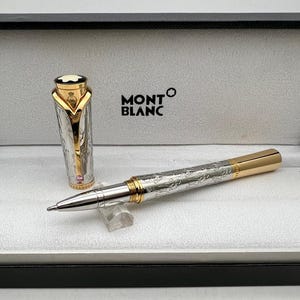 Puede incluir: Un bolígrafo rollerball plateado y dorado con intrincados detalles grabados, presentado en una caja negra con el logotipo de Montblanc. El capuchón del bolígrafo está chapado en oro y se muestra sobre una superficie blanca.
