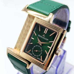 Puede incluir: Un reloj de lujo con una esfera verde rectangular y una caja dorada. El reloj presenta una correa de cuero verde y la marca "Jaeger-LeCoultre" en la esfera. El reloj se exhibe sobre un soporte transparente.