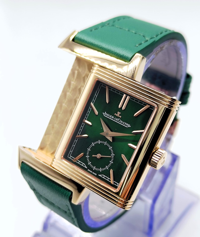 Puede incluir: Un reloj de lujo con una esfera verde rectangular y una caja dorada. El reloj presenta una correa de cuero verde y la marca "Jaeger-LeCoultre" en la esfera. El reloj se exhibe sobre un soporte transparente.