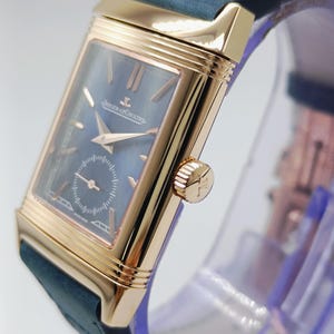 K&ouml;nnte beinhalten: Eine rechteckige, goldfarbene Armbanduhr mit dunkelblauem Zifferblatt und kleiner Sekunde. Die Uhr hat goldene Zeiger und Markierungen, eine Krone mit Logo und ein dunkelblaues Lederarmband. Die Uhr wird auf einem transparenten St&auml;nder pr&auml;sentiert.